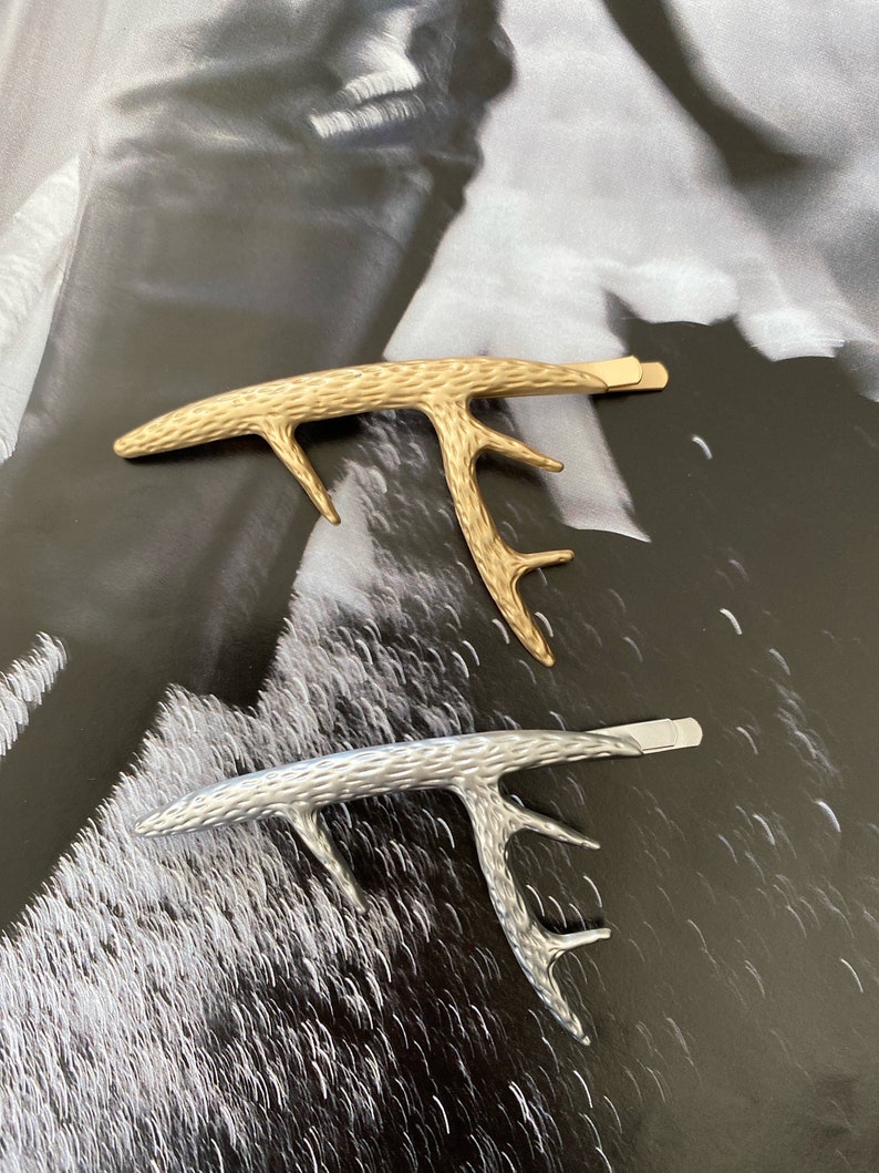 Peut inclure: Deux pinces &agrave; cheveux en forme de bois de cerf. L'une est dor&eacute;e, l'autre argent&eacute;e. Les deux pinces ont une surface textur&eacute;e et une fermeture de type barrette. Les pinces sont pr&eacute;sent&eacute;es sur un fond noir et blanc.