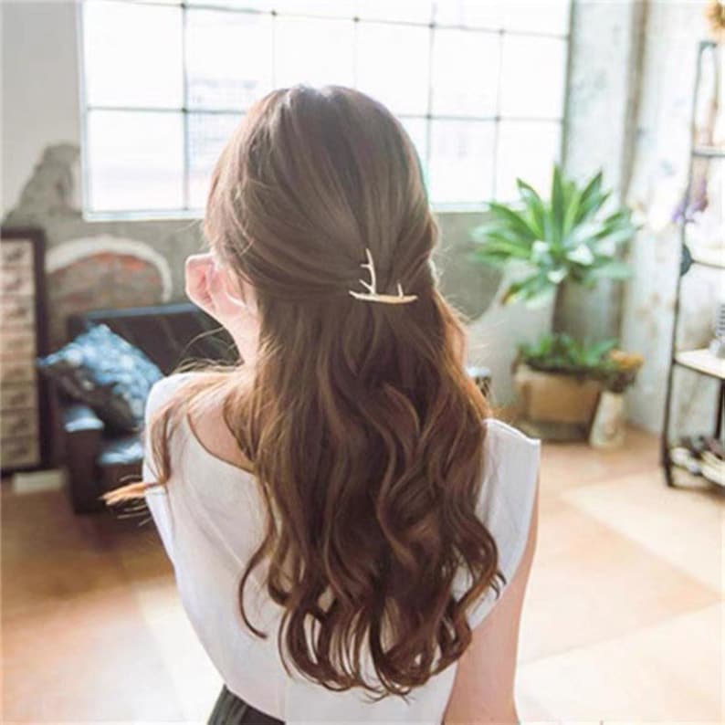 Peut inclure: Une femme aux cheveux bruns et ondul&eacute;s porte une pince &agrave; cheveux dor&eacute;e en forme de bois de cerf. La pince est plac&eacute;e sur le c&ocirc;t&eacute; droit de la t&ecirc;te, ajoutant une touche d&eacute;corative &agrave; la coiffure. La femme porte un haut blanc.