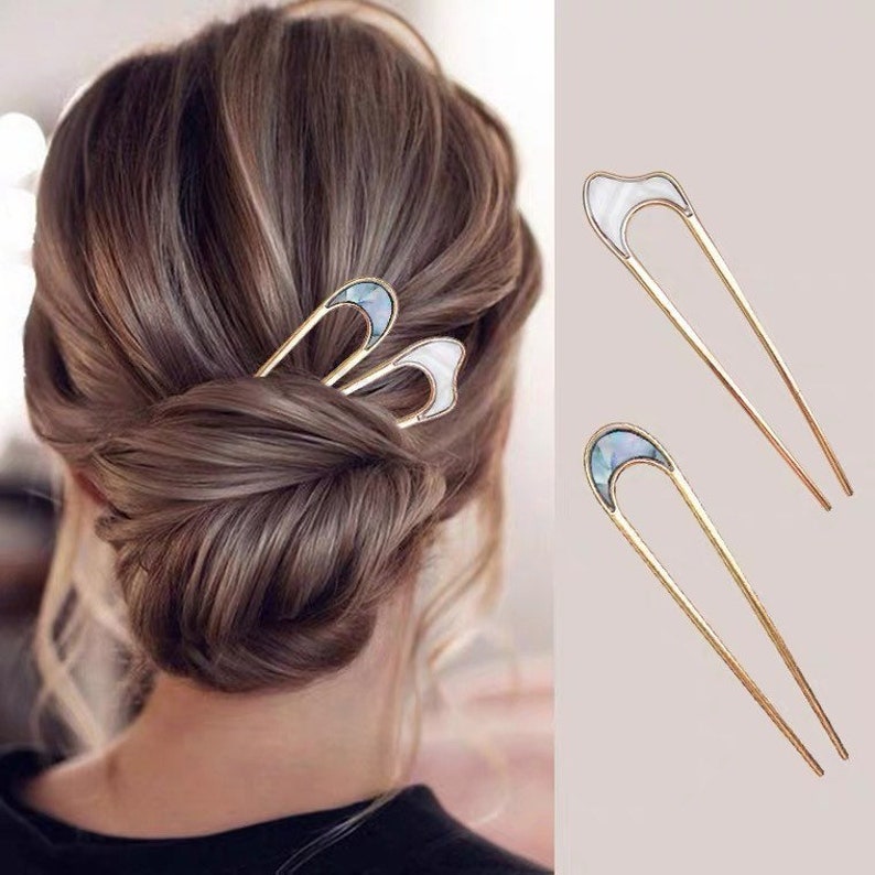 May include: Two gold hair pins with a white and blue iridescent shell design. The pins are shown in a close-up view and are being used to hold a woman's hair in a bun.