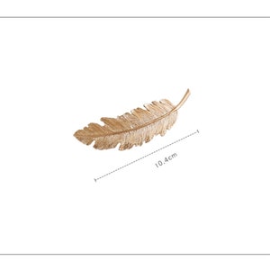 Peut inclure: Un objet d&eacute;coratif en forme de plume de couleur dor&eacute;e. La plume mesure 10,4 cm de long, avec des veines d&eacute;taill&eacute;es et une surface textur&eacute;e. L'objet est probablement en m&eacute;tal et est pos&eacute; sur un fond blanc.