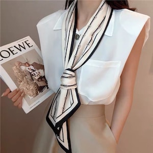 May include: A white and black patterned silk scarf with a geometric design. The scarf is tied around the neck and hangs down the front of the body.