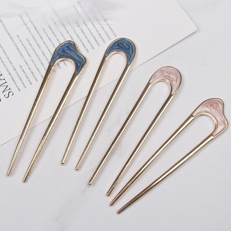 May include: Set of five gold hair sticks with decorative tops. Two sticks have blue tops with a swirl pattern, and three sticks have pink tops with a swirl pattern.