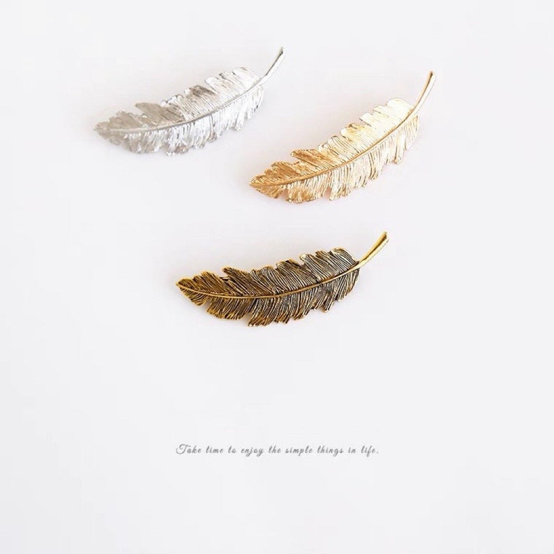 Peut inclure: Trois barrettes en m&eacute;tal en forme de plumes, argent&eacute;es, dor&eacute;es et bronze. Les plumes pr&eacute;sentent des veines d&eacute;taill&eacute;es et sont dispos&eacute;es sur un fond blanc. Le texte "Take time to enjoy the simple things in life" est en bas.