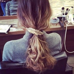Peut inclure: Une barrette en forme de plume dor&eacute;e attachant une queue de cheval basse. Les cheveux sont un m&eacute;lange de brun et de blond, l&eacute;g&egrave;rement ondul&eacute;s. La personne porte un pull &agrave; col roul&eacute; gris. Le fond montre un miroir et un salon.