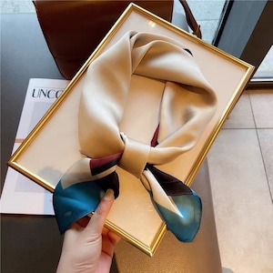 May include: A beige silk scarf with a geometric pattern in shades of blue, black, and red. The scarf is tied in a knot and is displayed in a gold frame.