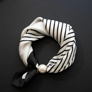 May include: A silk scarf with a black and white striped pattern and a solid black and white section. The scarf is knotted and secured with a large, round, pearl-like clasp. The scarf is on a black surface.