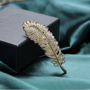 Peut inclure: Une broche en forme de plume dor&eacute;e orn&eacute;e de nombreuses petites perles blanches rondes. La broche est pos&eacute;e sur un fond sombre et un tissu de couleur sarcelle, soulignant son design complexe et son attrait &eacute;l&eacute;gant. Les d&eacute;tails de la plume sont finement travaill&eacute;s.