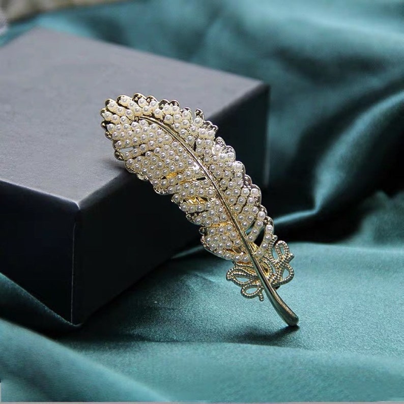 Peut inclure: Une broche en forme de plume dor&eacute;e orn&eacute;e de nombreuses petites perles blanches rondes. La broche est pos&eacute;e sur un fond sombre et un tissu de couleur sarcelle, soulignant son design complexe et son attrait &eacute;l&eacute;gant. Les d&eacute;tails de la plume sont finement travaill&eacute;s.