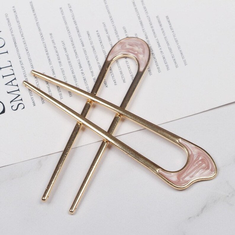 May include: A pair of gold hair chopsticks with a pink and white swirl design on the top. The chopsticks are about 6 inches long.