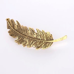 Peut inclure: Une barrette &agrave; cheveux en forme de plume en m&eacute;tal dor&eacute;. La barrette repr&eacute;sente une plume avec des nervures d&eacute;taill&eacute;es. La plume mesure environ 10 cm de long et poss&egrave;de une attache de type barrette &agrave; l'arri&egrave;re.