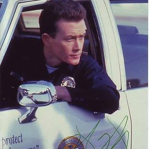 May include: A man in a black jacket and a badge is sitting in a white police car. The car has the words "protect and to serve" written on the side. The car also has a Los Angeles Police Department seal on the side. The man is looking to the right of the image.