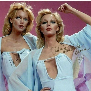 May include: Two women in light blue robes with plunging necklines. The women are smiling and looking at the camera. The robes are sheer and have a silky texture. The women are both wearing their hair long and flowing. The background is a light pink.