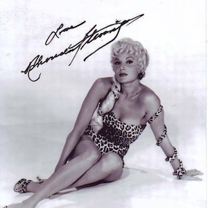 May include: Black and white photo of a woman wearing a leopard print swimsuit and a snake-like arm band. She is sitting with her legs crossed and her right hand resting on the floor. The woman has blonde hair and is wearing a small earring in her right ear. The photo is signed "Love,  Ann-Margret".