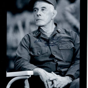 May include: A black and white photo of a man wearing a military uniform and a cap. He is sitting in a chair and has his hands clasped in his lap. He has signed his name in blue ink on the photo. The signature reads "All the best!"