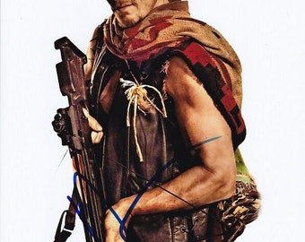 May include: A person wearing a leather vest and a patterned scarf, holding a crossbow. The person has a serious expression and is looking towards the camera. The background is plain white.