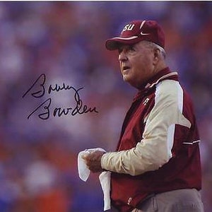May include: A black and white autograph of Bobby Bowden, a former American football coach. The signature is on a white background. Bowden is wearing a maroon and gold jacket with a maroon hat that says "FSU" on it.