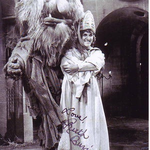 May include: A black and white photo of a woman in a white dress and a crown, standing next to a large, furry, monster-like creature. The woman is smiling and the creature has a large, toothy grin. The woman's autograph is visible on the dress, "Love, Ruth Buzzi."