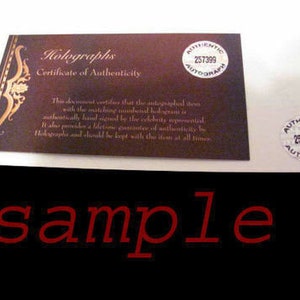 May include: A certificate of authenticity with a brown background and gold accents. The certificate states that the autographed item is authentic and includes a matching numbered hologram. The certificate also includes the text "Holographs Certificate of Authenticity" and "Authentic Autograph 257399".