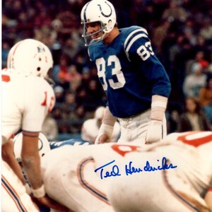 May include: A football player wearing a blue jersey with the number 83 on it. The player is wearing a white helmet with a blue horseshoe logo. The player is standing on a football field and is looking down at the ground. The player's signature, "Ted Hendricks", is written in blue ink below the image.
