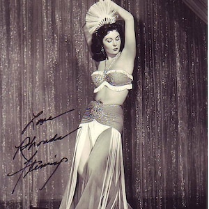 May include: A black and white photo of a woman wearing a white costume with fringe and a feathered headdress. She is posing with her arms raised above her head. The photo is signed "Love Rhonda Fleming".
