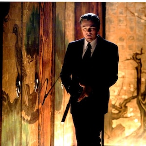 May include: A man in a black suit and tie stands in a room with a dark wood panelled wall. The wall is decorated with a large painting of a bird. The man is looking to the right of the image.