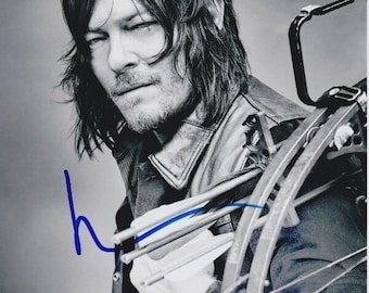May include: Black and white photo of a man with long hair holding a crossbow. The man is looking at the camera with a serious expression. The crossbow is in focus and the man is out of focus.