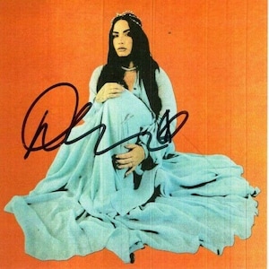 May include: A woman with long dark hair wearing a light blue gown with a crown on her head. The woman is sitting on an orange background and has a signature in black ink.