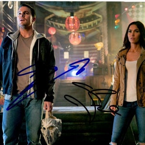 May include: A man and a woman in a dark, industrial setting. The man is wearing a blue denim jacket and jeans, and is holding a mask. The woman is wearing a brown jacket and jeans.