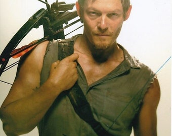 May include: A man with short brown hair is wearing a grey sleeveless shirt and holding a black crossbow on his shoulder. He is looking at the camera with a serious expression. The crossbow has a red arrow in the bow.