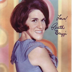 May include: A woman with short, dark brown hair is wearing a light blue sleeveless top. She is smiling and looking to the right. The background is a colorful abstract design. The image is signed "Love! Ruth Buzzi".