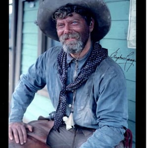 May include: A man wearing a blue shirt and a brown cowboy hat with a black and white bandana around his neck. He is sitting on a wooden porch with a wooden door behind him. The man has a beard and mustache and is looking to the side. The image is signed by the man.
