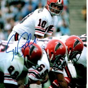 May include: A football player wearing a white jersey with black and red stripes, a red helmet with a white falcon logo, and the number "10" on the jersey. The player is in a football huddle with other players wearing red helmets and white jerseys with black and red stripes.