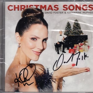 May include: CD case for "Christmas Songs" by David Foster & Katharine McPhee. The cover shows a woman with a bun and a man playing piano in a snow globe. The CD case is signed by the artists.