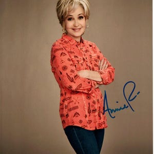 May include: A photograph of a woman with short, blonde hair, wearing an orange button-down shirt with a Southwestern pattern and dark blue jeans. The shirt is adorned with black cacti and geometric designs. The image includes a signature in blue ink.
