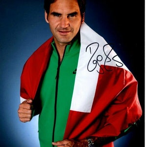 May include: A man wearing a green, white, and red jacket with a white stripe down the middle. He is holding the jacket up to his shoulders like a flag. The jacket is signed "Roger Federer".