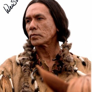 May include: A close-up portrait of a man with long dark hair wearing a brown leather jacket with fur trim. The man is looking to the side and has a serious expression on his face. The image is signed "Wes Studi" and dated "2018".
