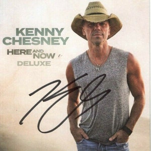 May include: A signed album cover featuring Kenny Chesney. The album is titled "Here and Now Deluxe". The cover features a photo of Kenny Chesney wearing a grey tank top, blue jeans, and a brown cowboy hat. He is looking at the camera and smiling.