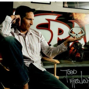 May include: A man wearing a striped shirt and jeans is sitting in a chair. He is holding a small skull-shaped object in his hand. The background features a large graphic of the word "Spawn" with a red and white color scheme. The man has signed his name "Todd McFarlane" in silver marker.