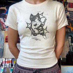 Könnte beinhalten: Cremefarbenes T-Shirt mit einem schwarz-weißen Motiv einer Katze, die Gitarre spielt, umgeben von Sternen. Das kurzärmelige Shirt ist aus weichem Material.