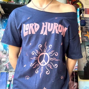 Può includere: T-shirt blu navy con spalle scoperte con la scritta "LORD HURON" in rosa, sopra un disegno di sole rosa con un simbolo di pace al centro. La maglietta presenta anche piccole stelle rosa.