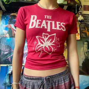 以下が含まれることがあります： 白い花の模様の上に、白い文字で「The Beatles」と書かれた赤いTシャツ。半袖のシャツは柔らかい素材で作られています。