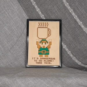 May include: Framed artwork featuring a pixelated character holding a coffee mug. The character is in shades of brown and green. The text "IT'S DANGEROUS TO GO ALONE! TAKE THIS." is below the character. The frame is black.