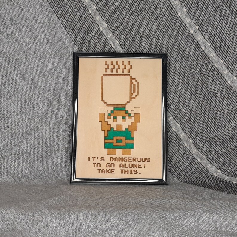 May include: Framed artwork featuring a pixelated character holding a coffee mug. The character is in shades of brown and green. The text "IT'S DANGEROUS TO GO ALONE! TAKE THIS." is below the character. The frame is black.