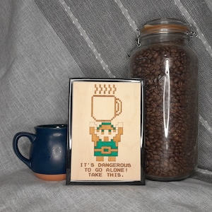 May include: A framed wooden art piece with a pixelated character holding a coffee mug, with the text "IT'S DANGEROUS TO GO ALONE! TAKE THIS." A blue mug and a jar of coffee beans are also in the image.