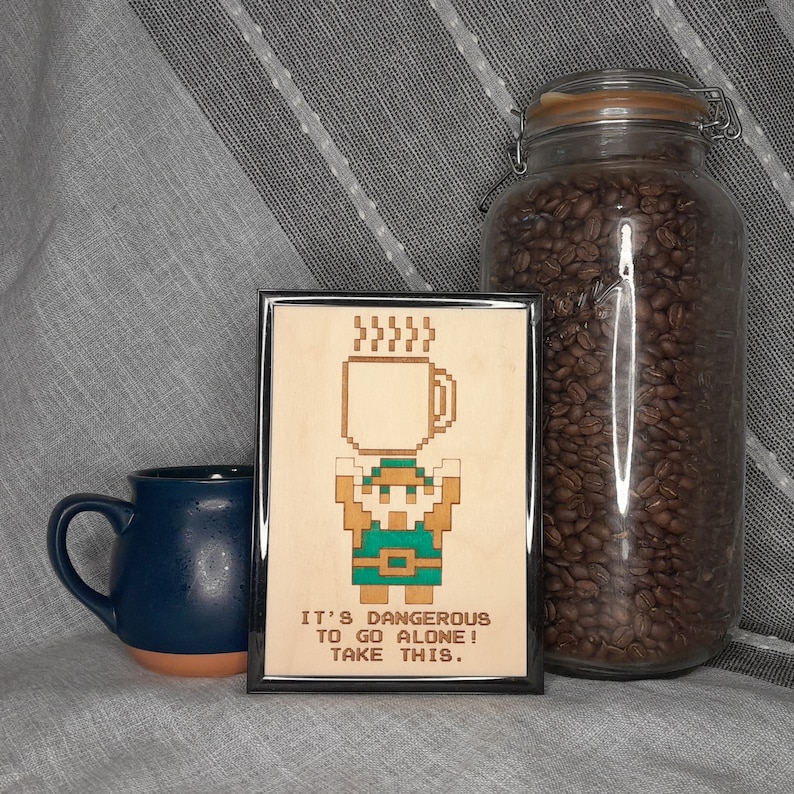 May include: A framed wooden art piece with a pixelated character holding a coffee mug, with the text "IT'S DANGEROUS TO GO ALONE! TAKE THIS." A blue mug and a jar of coffee beans are also in the image.