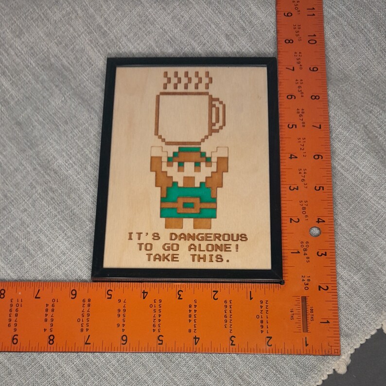 May include: Wooden framed art piece featuring a pixelated character holding a coffee mug. The artwork is in shades of brown and green, with the text "IT'S DANGEROUS TO GO ALONE! TAKE THIS." The frame is black.