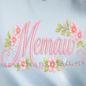 May include: Light blue t-shirt with the embroidered word "Memaw" in pink script, surrounded by pink flowers and green leaves. The shirt has rolled-up sleeves and a crew neck. The text "Memaw Designs" is visible at the bottom.