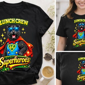 Puede incluir: Camiseta negra con un dise&ntilde;o de pantera de dibujos animados con gafas de sol, capa roja y disfraz de superh&eacute;roe. La camiseta tiene el texto "Lunch Crew Superheroes" en amarillo y verde. El dise&ntilde;o incluye estrellas amarillas.