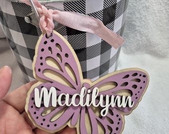 May include: A wooden butterfly cutout with a purple paint finish and the name "Madilynn" written in white. The butterfly is attached to a pink ribbon and a black and white checkered metal bucket.