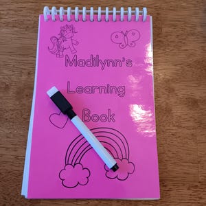 May include: Pink learning book with a spiral binding. The cover features a unicorn, a butterfly, a rainbow, and clouds. The book has the text "Madilynn's Learning Book" on the cover. A white dry-erase marker is lying on top of the book.